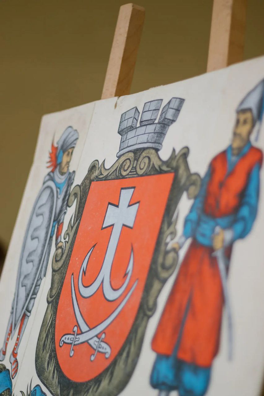 Великий герб Вінниці: щит, на якому зображено хрест з гачками та шаблі на червоному полі, його тримають козак та лицар